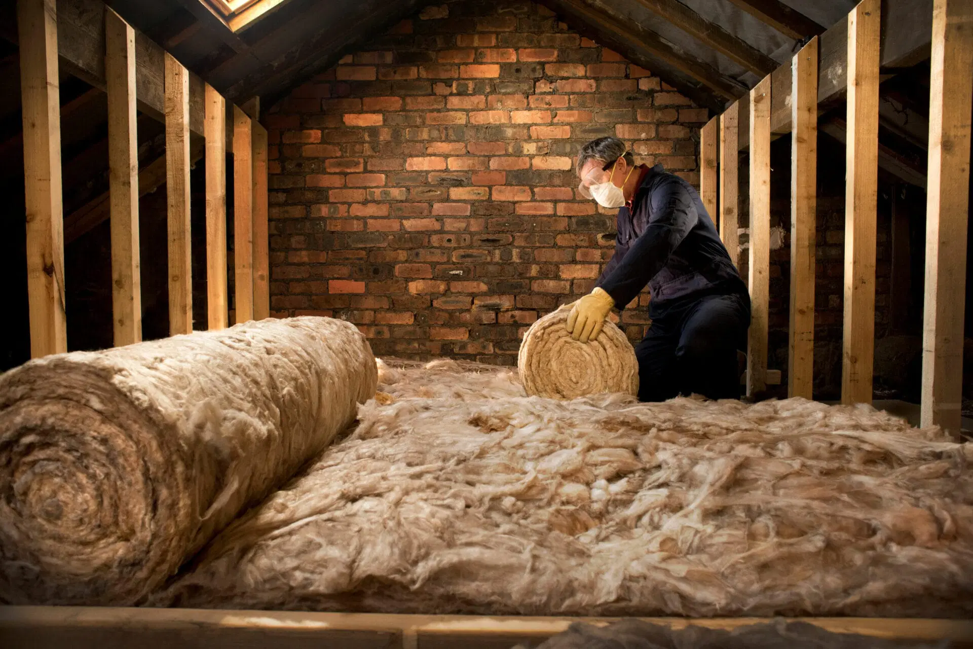 Worker installing roll insulation in attic space with wooden framing and brick wall background 