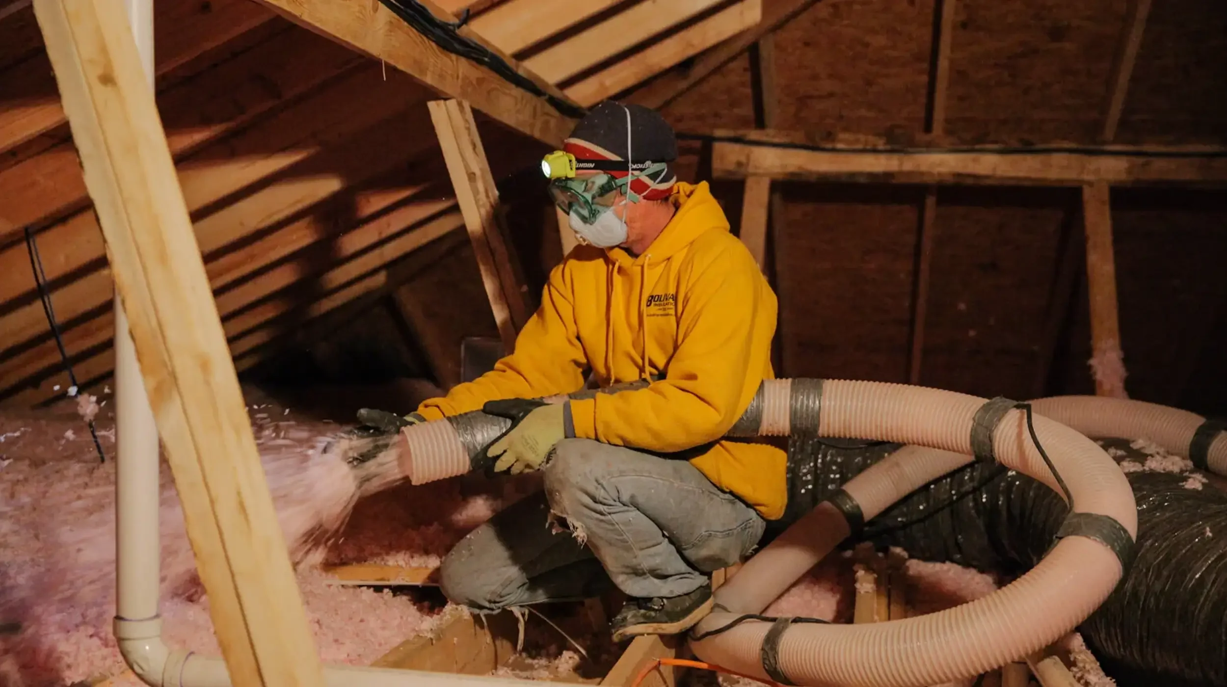 Close-up of a worker operating a large blower hose to apply pink loose-fill insulation between roof trusses