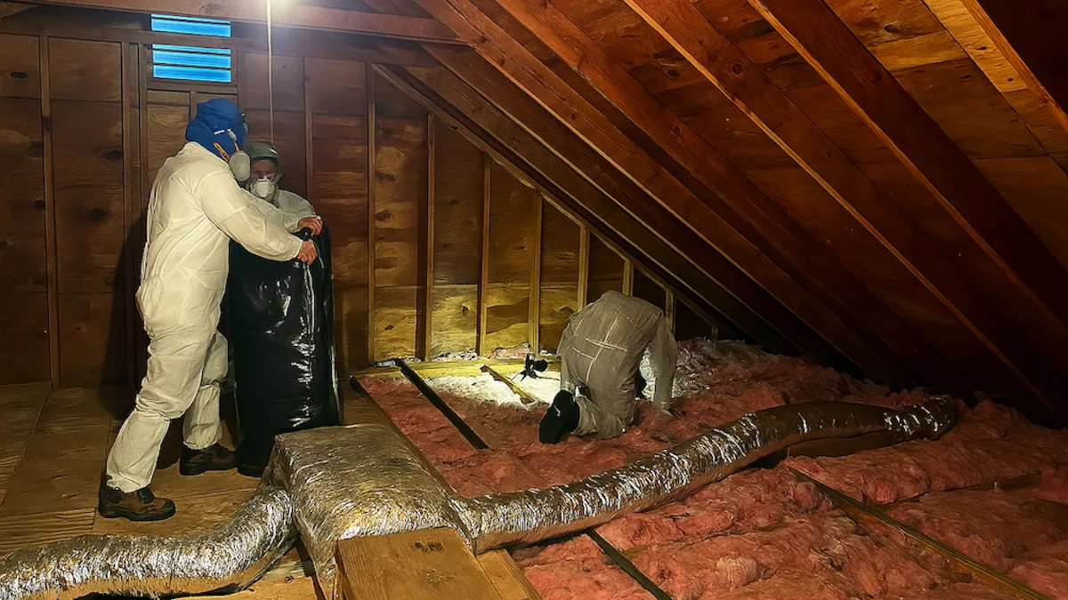 Insulation removal technicians wearing respirators and suit protection filling heavy-duty waste bags in an attic